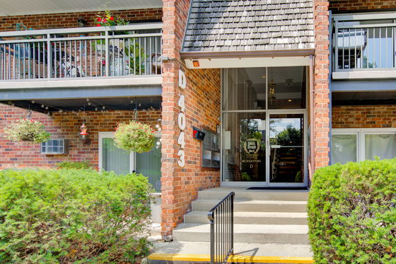 Front view of a brick building entrance with a balcony and hanging plants.
