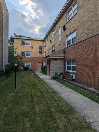 Front view of a multi-story brick apartment building with a walkway and grass lawn.