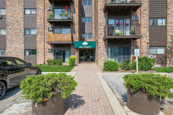Front view of a multi-story brick apartment building with balconies and a green awning over the entrance.