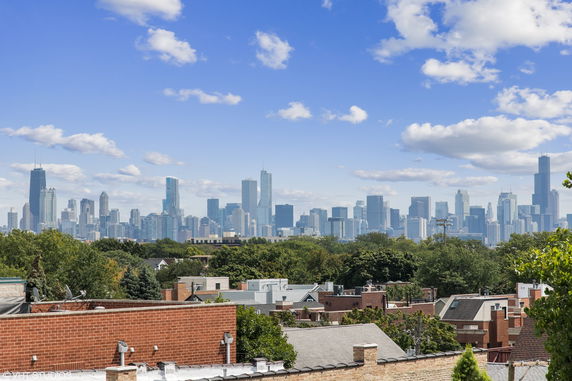 Panoramic view of a city skyline from a distance, with a variety of tall buildings against a blue sky and scattered clouds.