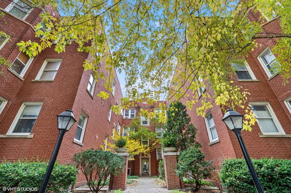 Front view of a brick apartment building with trees and a pathway leading to the entrance.