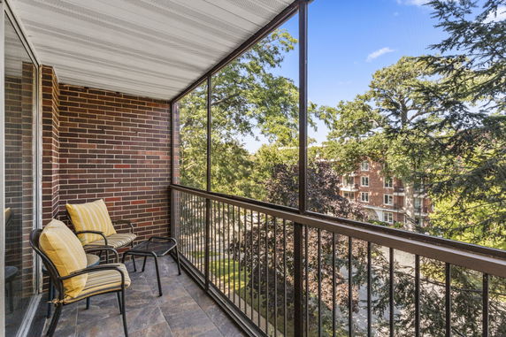 Outdoor balcony with chairs and railing, overlooking trees and a building in the background.