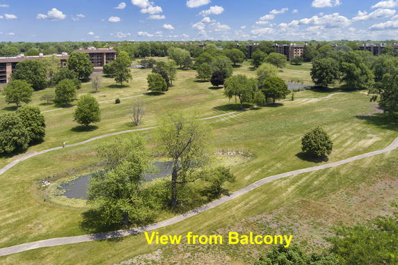 Wide view of a grassy landscape with scattered trees and buildings in the distance, as seen from a balcony.
