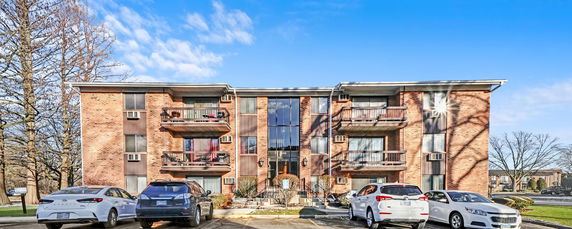 Front view of a three-story residential building with balconies and cars parked in front.