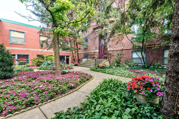Front view of a brick building entrance with lush gardens and colorful flowers.