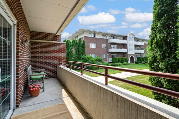 View from a balcony showing a brick apartment building with multiple windows and balconies, surrounded by trees and greenery.