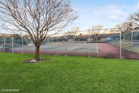 Outdoor tennis court surrounded by a wire fence, with a tree and a bench nearby.
