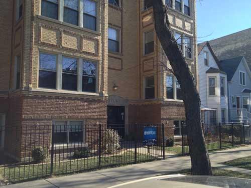 Front view of a multi-story brick apartment building with a wrought iron fence.