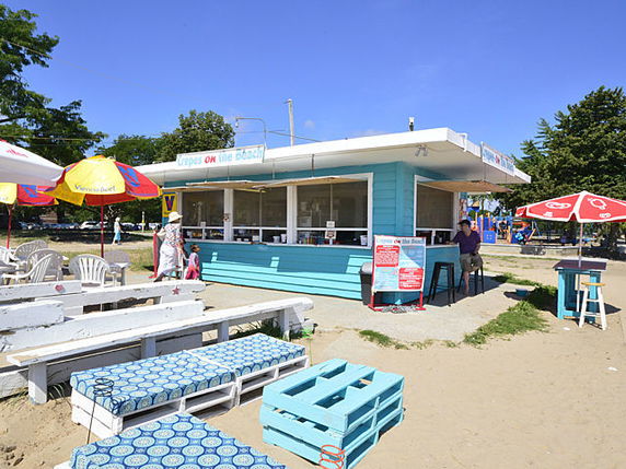 Front view of a beachside kiosk with a flat roof and large windows, offering a casual seating area with colorful umbrellas.