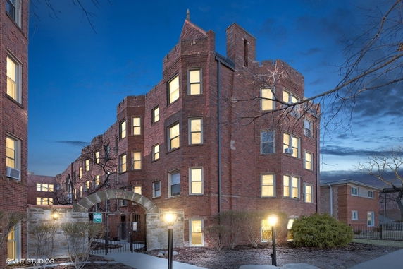 Front view of a brick multi-story building with illuminated windows and an arched entrance.