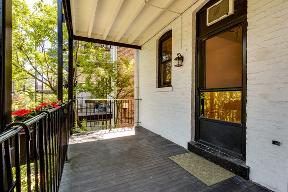 View of a building entrance with a porch, featuring a black door, railing, and shaded area under a canopy.