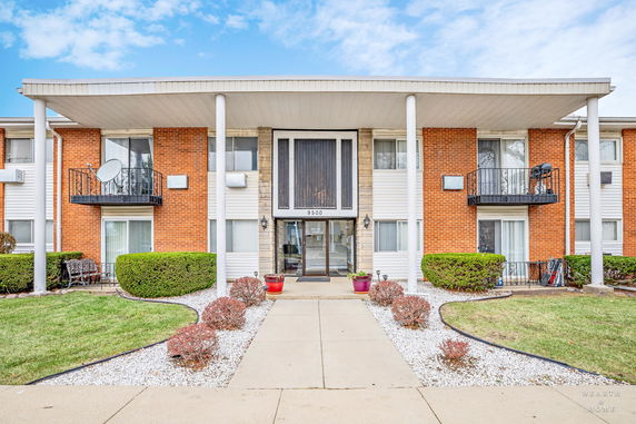Front view of a two-story residential building with brick and panel siding, featuring balconies and entry columns.