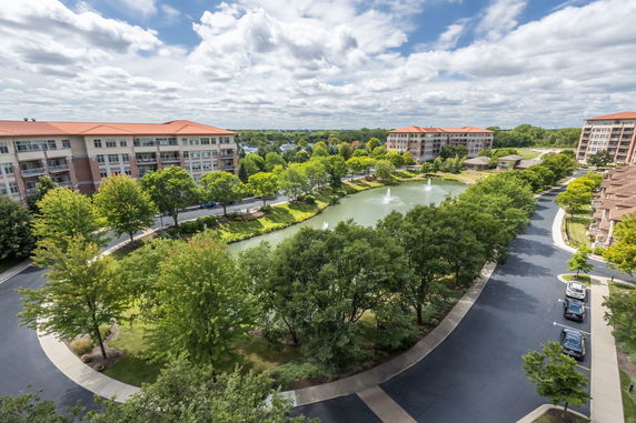 Panoramic view of multi-story buildings with a central pond and fountain, surrounded by trees and roads.
