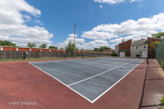 Tennis court surrounded by a fence with a building in the background.