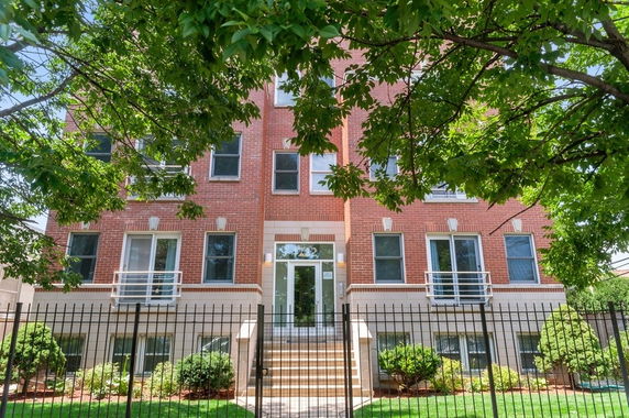Front view of a multi-story brick building with balconies and a fenced entrance.