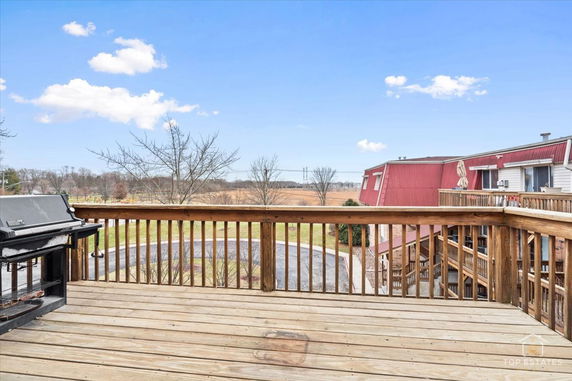 Wooden deck with railing offering a view of surrounding open field and neighboring building rooftops.