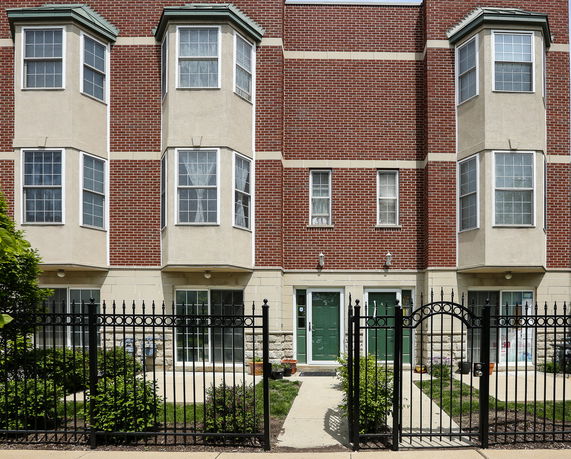 Front view of a multi-story brick townhouse with bay windows and green doors behind a black metal fence.