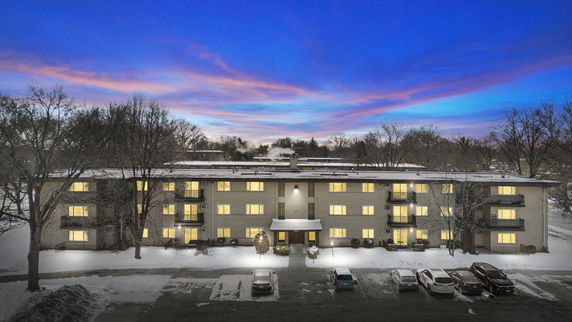 Front view of a multi-story residential building with lit windows and balconies, surrounded by snow and parked cars.