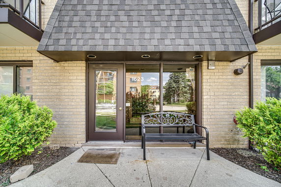 Front view of a building entrance with a glass door and windows, sheltered by a shingled overhang.