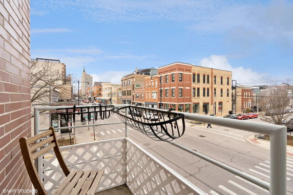 Panoramic view of an urban street with buildings and a balcony in the foreground.