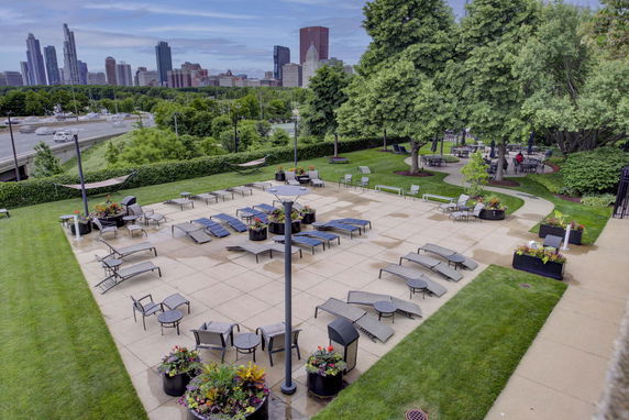 Rooftop leisure area with lounge chairs and tables, surrounded by greenery and trees, with a city skyline in the background.