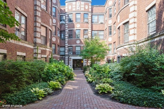 Front view of a multi-story brick apartment building with an entrance pathway and greenery.