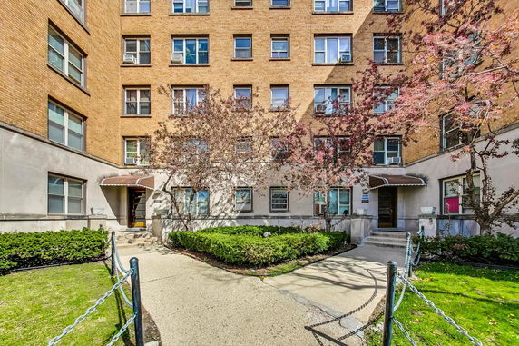 Front view of a multi-story brick apartment building with a central entrance and surrounding landscaping.
