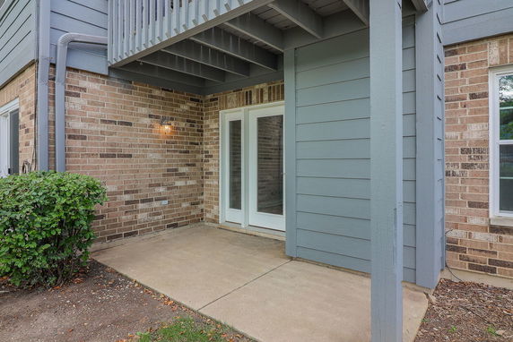 Front view of a building entrance with brick and siding walls, featuring a patio area and double glass doors.