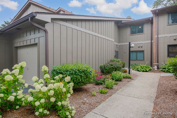 Front view of a two-story building with siding and a garage door, surrounded by landscaped plants and a walkway.