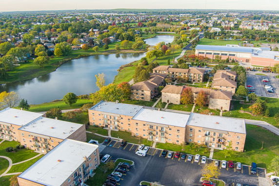 Aerial panoramic view of residential buildings, surrounding greenery, and a nearby lake.