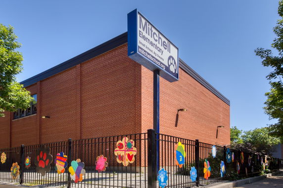 Front view of a brick elementary school building with a sign and colorful decorations on the fence.