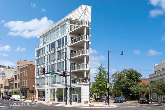 Front view of a modern multi-story building with glass balcony railings and a unique slanted roof design.