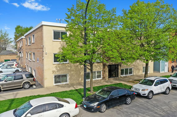 Front view of a three-story brick condominium building with trees and parked cars in front.