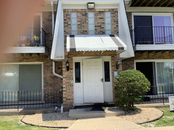 Front view of a brick apartment building entrance with a white door and awning, featuring balconies on either side.