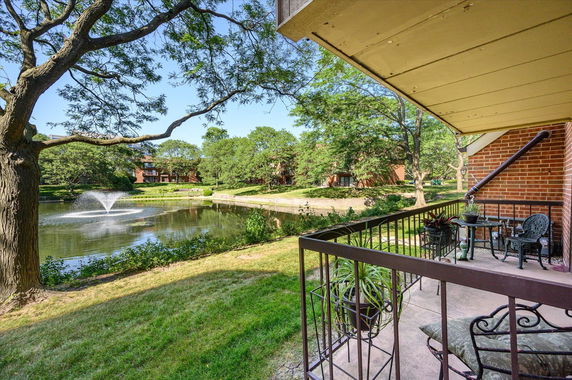 View of a pond with a fountain surrounded by trees and apartment buildings, seen from a patio with railing and outdoor furniture.