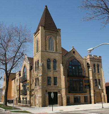 Front view of a brick church-style building with a tall spire and large arched windows.