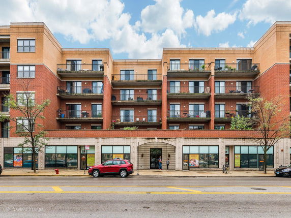 Front view of a multi-story brick building with balconies and ground-level retail spaces.
