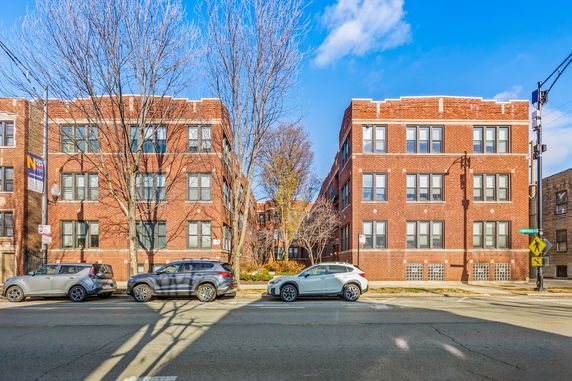 Front view of a brick-finished apartment building with multiple windows and parked cars on the street.
