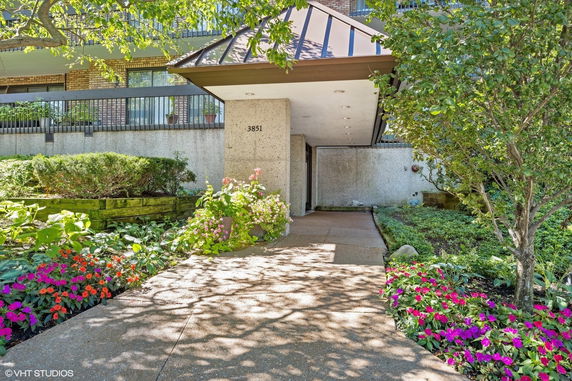 Front view of a building with a covered entrance walkway, surrounded by landscaping and flowers.