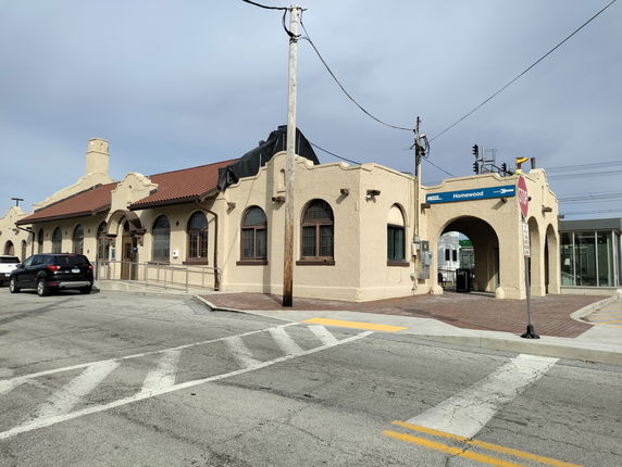 Front view of a tan-colored building with a red tile roof and arched windows, identified as a station with signs.