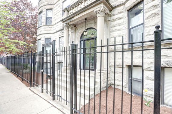 Front view of a stone building with ornate columns and windows, surrounded by a black metal fence.