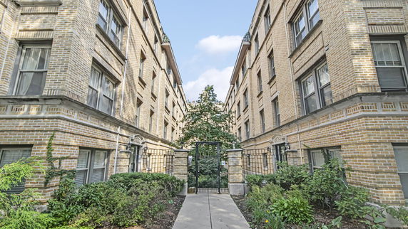 Front view of a multi-story brick building with a gated entrance and greenery on both sides of the pathway.