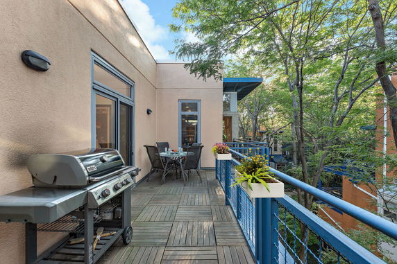 Balcony area with a grill and dining setup, surrounded by trees and adjacent building.