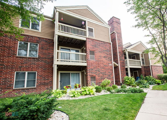 Front view of a multi-story building with brick and wood siding, featuring balconies and a landscaped area.