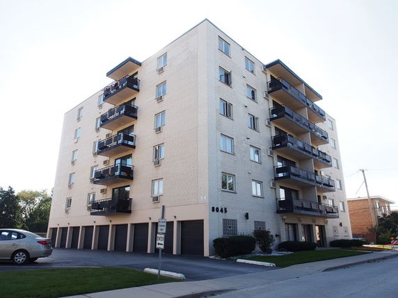 Front view of a multi-story apartment building with balconies and a parking area.