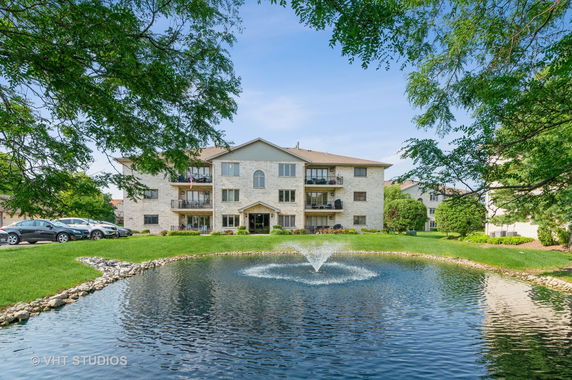 Front view of a multi-story building with balconies and a small pond with a fountain in front.
