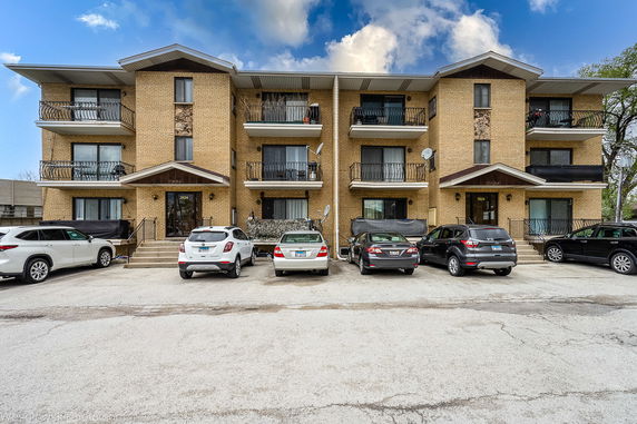 Front view of a brick apartment building with balconies and a parking area in front.
