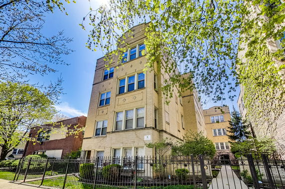 Front view of a multi-story apartment building with beige brickwork and a metal fence.