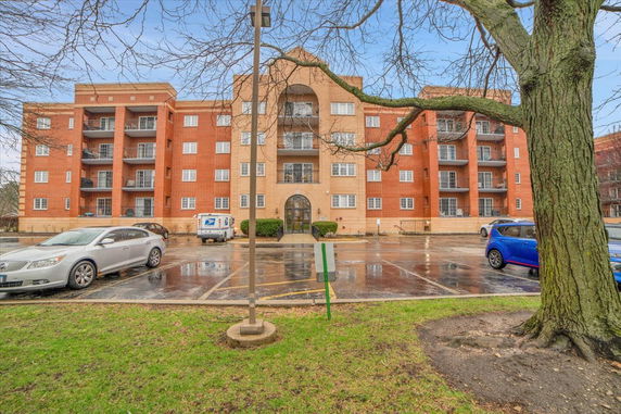 Front view of a multi-story brick apartment building with balconies and surrounding parking area.