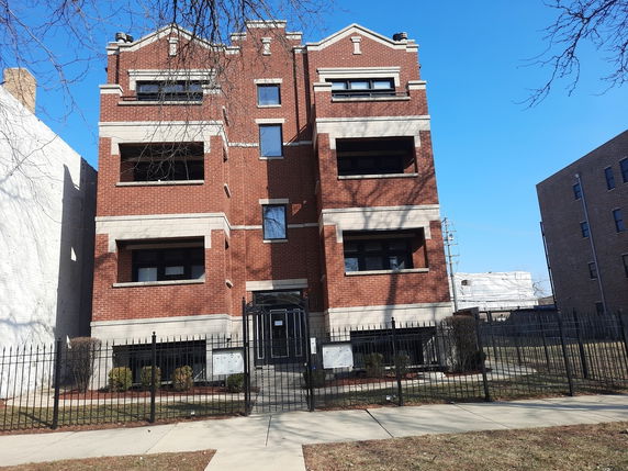 Front view of a three-story red brick apartment building with balconies and a gated entrance.
